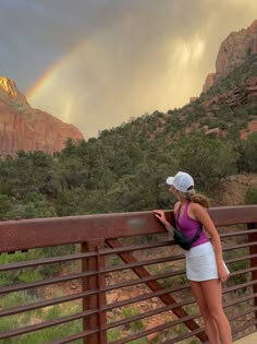 a woman is standing on a bridge looking at the mountains with a rainbow in the sky