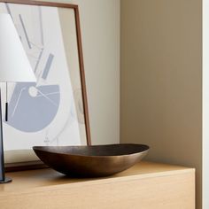 a wooden bowl sitting on top of a dresser next to a lamp and framed photograph