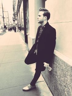 a man in a suit leaning against a wall on the side of a building with his hand under his leg