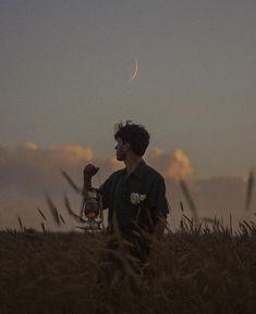 a man standing in tall grass holding a lantern and looking up at the sky with a half moon behind him