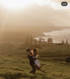 a man and woman standing on top of a lush green hillside next to the ocean