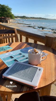 a tablet sitting on top of a wooden table next to the ocean with a cup of coffee