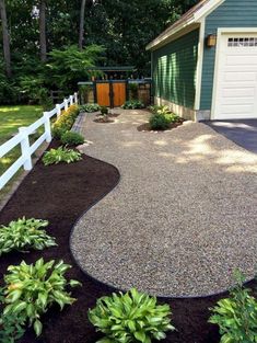 a driveway with gravel and plants in front of the garage, next to a white picket fence