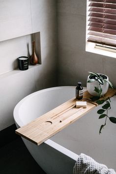 a white bath tub sitting under a window next to a wooden tray with a plant on it