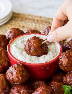 a hand dipping some meatballs into a small red bowl with cream cheese on top