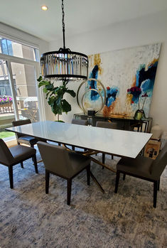 a dining room table and chairs in front of a large window with a painting on the wall