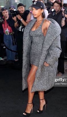 a woman in a grey coat and heels standing on the black carpet with cameras around her