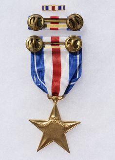 Back side view of the Silver Star medal, which is the third-highest military decoration for valor awarded to members of the United States Armed Forces. Any uniformed service member may receive the medal, which is awarded for gallantry in action against an enemy of the United States. Also shown in the ribbon lapel bar and the back side of the ribbon bar, which features a double clutch back bar.  The reverse has the inscription FOR GALLANTRY IN ACTION. Back Bar, Side View, Arrow Necklace