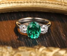 an emerald and diamond ring on top of a wooden table next to a gold frame