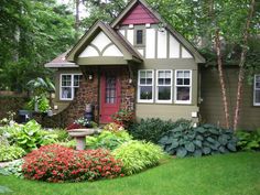 a small house surrounded by lush green grass and flowers in the front yard with red door