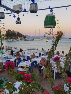 many people are sitting at tables near the water with flowers in front of them and lights hanging from the ceiling