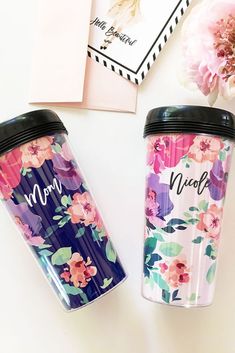 two travel mugs sitting next to each other on top of a white table with pink flowers