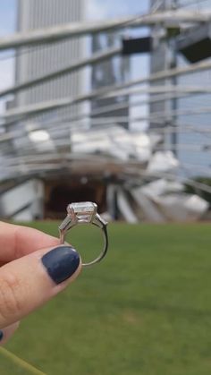 a hand holding a ring with a diamond on it in front of some grass and buildings