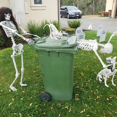 two skeletons are standing next to a trash can in the grass, and one skeleton is sitting on it's side