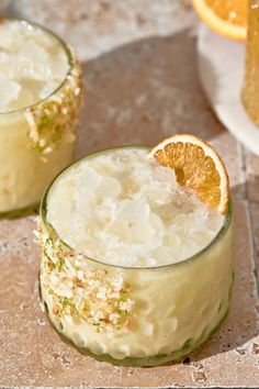 two glasses filled with ice and oranges sitting on a tile counter top next to each other