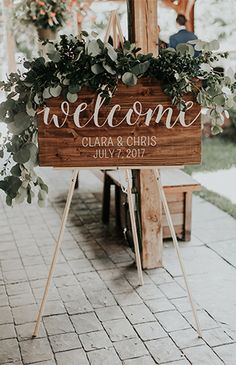 a wooden welcome sign sitting on top of a brick floor next to a table with greenery