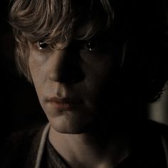 a young man with blonde hair and dark eyes stares at the camera in a dimly lit room