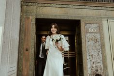 a woman in a white dress is walking out of a building with a man wearing a suit and tie