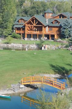 a wooden bridge over a small pond in front of a large log house with a green roof