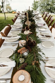 a long table set with plates, silverware and pine cones is decorated with greenery