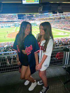 two young women standing next to each other in front of a crowd at a baseball game