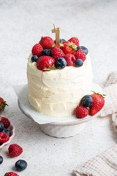 a white cake with berries and blueberries on top is sitting next to a bowl of strawberries