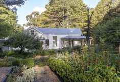 a small white house surrounded by trees and plants in the foreground is a garden