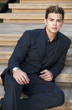 a man in a black suit sitting on some steps and looking at the camera with his hand on his hip