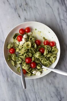 a white bowl filled with pesto pasta and cherry tomatoes on top of a marble table