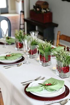 the table is set for christmas dinner with green napkins and greenery in glass vases