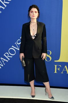 a woman standing in front of a blue wall wearing a black suit and matching heels