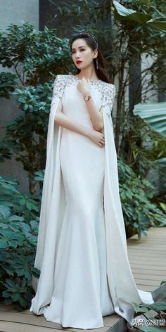 a woman in a white gown and cape standing on a wooden deck surrounded by greenery