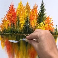 a hand is holding a paintbrush in front of a painting of colorful trees