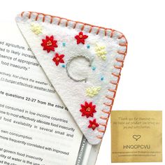 an open book with some red and white flowers on it next to a small triangle shaped brooch