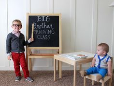 a little boy standing in front of a blackboard next to a small table and chair