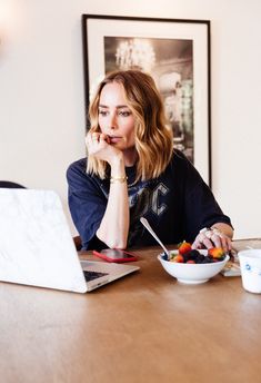 a woman sitting at a table with a bowl of fruit in front of her laptop