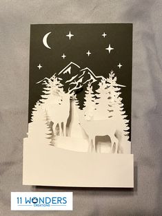 a card with two deer standing in front of trees and the moon above them, on a sheet of white paper