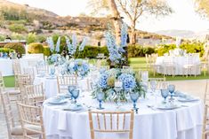 a table set up with blue flowers and white linens for an outdoor wedding reception