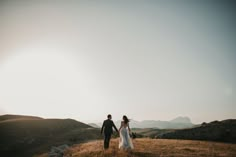 a bride and groom holding hands while walking through the grass on top of a hill