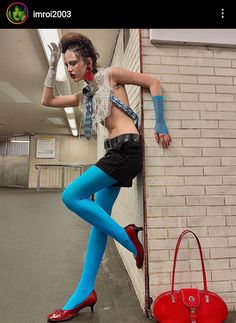 a woman in blue tights and heels leaning against a brick wall with a red purse