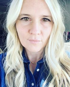 a woman with long blonde hair wearing a blue shirt and looking at the camera while sitting in a car