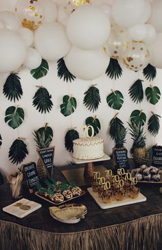 a table topped with lots of desserts next to white balloons and palm leaf decorations