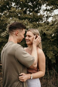 a man and woman standing next to each other in front of some trees with their arms around each other