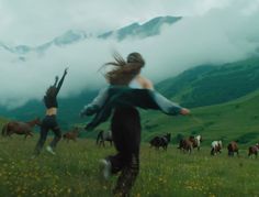 two women are running through a field with horses in the backgroung behind them