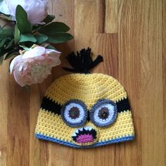 a crocheted minion hat next to flowers on a wooden floor with pink peonies