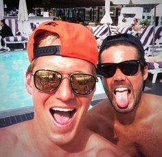 two men in sunglasses and an orange hat pose for the camera near a swimming pool