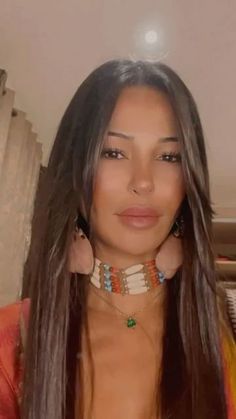 a woman with long hair wearing jewelry and looking at the camera while standing in front of a mirror