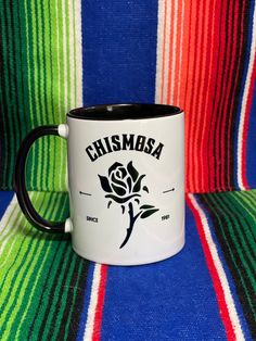 a black and white coffee mug sitting on top of a colorful striped blanket with the words, chimaga