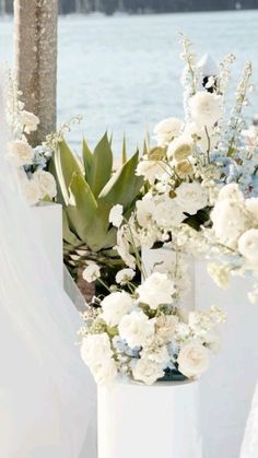 three white vases with flowers and greenery on the side of a body of water