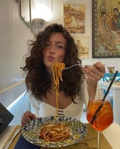 a woman sitting at a table eating spaghetti from a plate with a drink in front of her